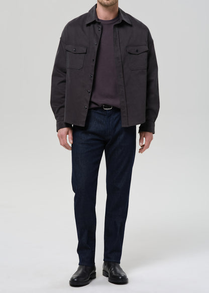 A man wearing a dark charcoal grey overshirt over a dark purple t-shirt, dark blue jeans, and black leather shoes, standing against a grey background.