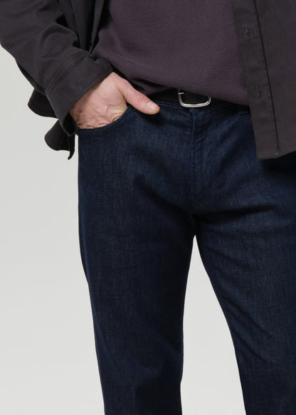 Close-up of a person wearing dark blue denim jeans with a silver belt buckle, a dark ribbed shirt, and a black jacket, with one hand in the pocket, against a grey background.