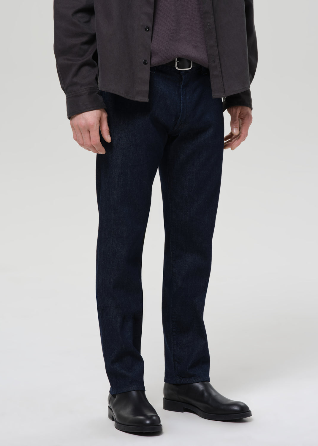 A person wearing dark navy blue jeans, a dark charcoal jacket, a dark shirt, and black leather shoes, standing against a grey background.