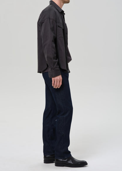 A man shown in profile wearing a dark charcoal button-up shirt, dark blue jeans, and black leather boots, photographed against a grey background.