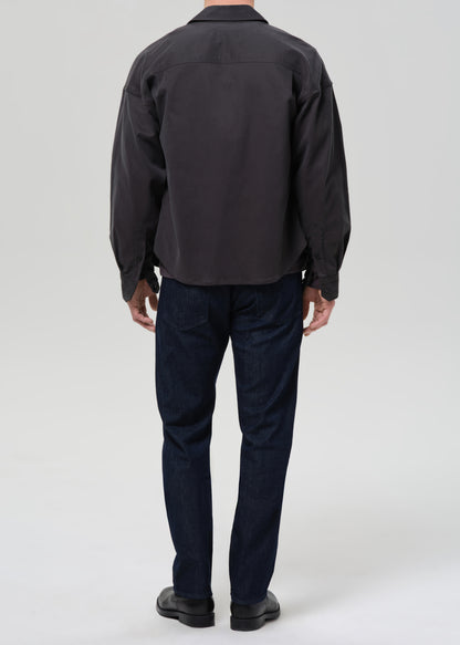 A man viewed from the back wearing a dark charcoal grey long-sleeve button-up shirt, dark blue jeans, and black shoes, standing against a grey background.