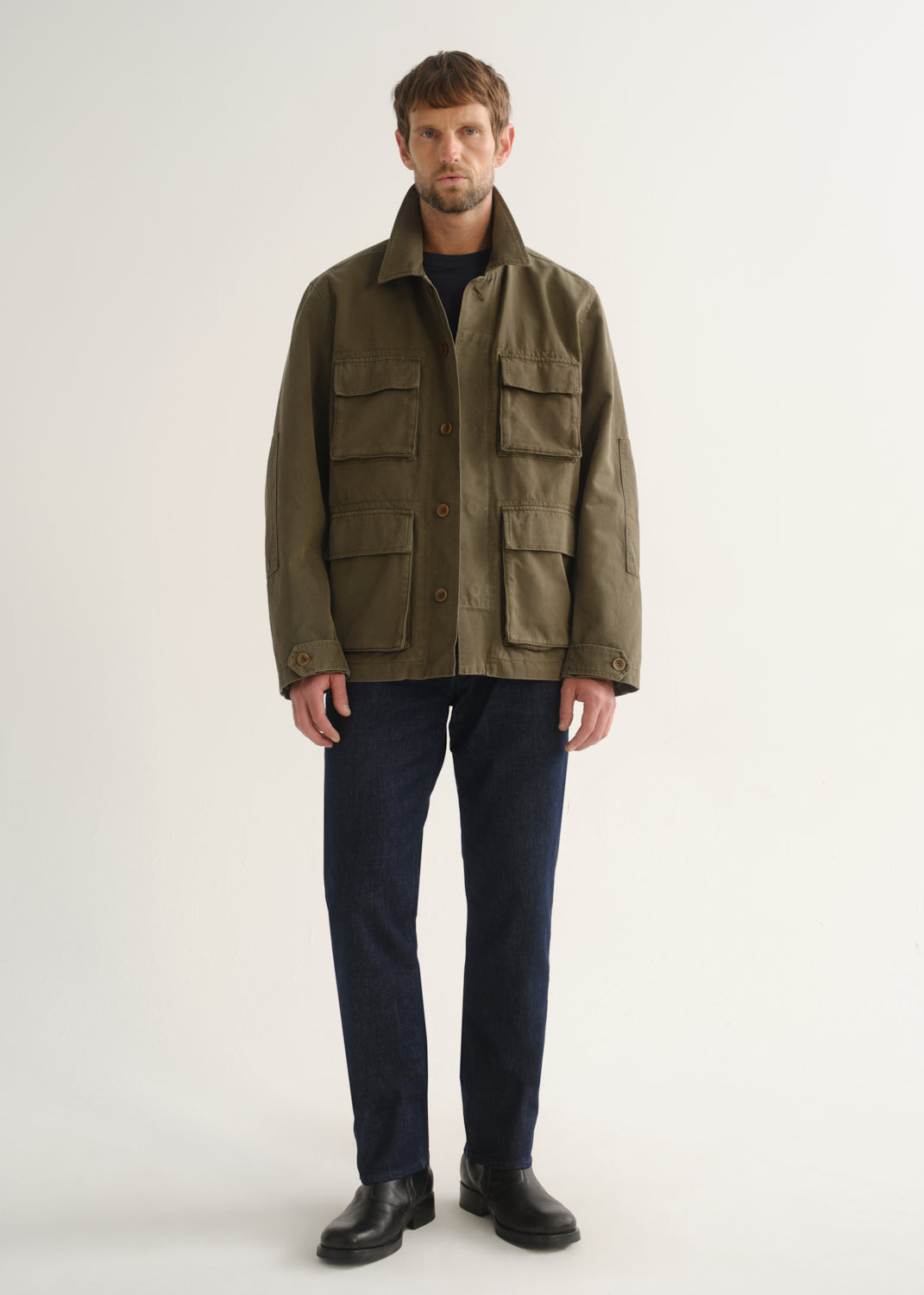 Person wearing dark indigo straight leg jeans with an olive green jacket and black boots, front full body view on a light grey background.