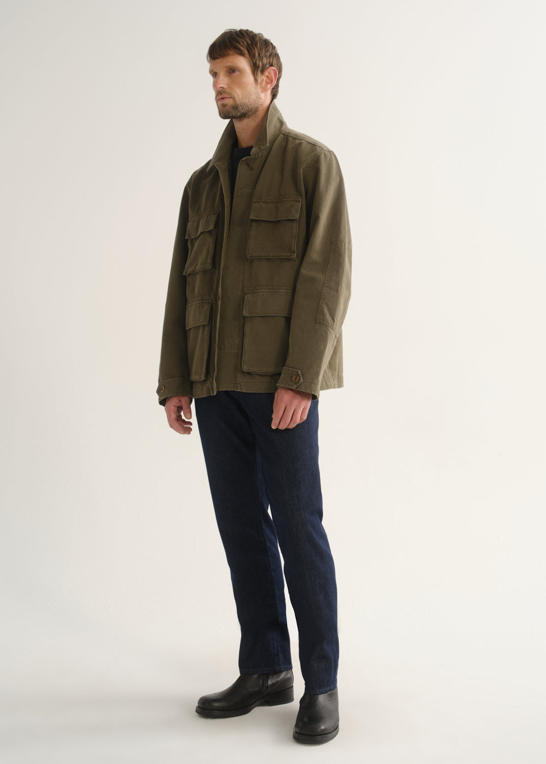 Person wearing dark indigo straight leg jeans with an olive green jacket and black boots, angled front full body view on a light grey background.