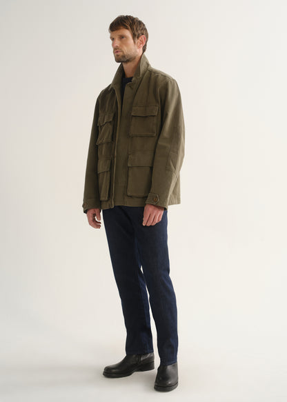 Person wearing dark indigo straight leg jeans with an olive green jacket and black boots, angled front full body view on a light grey background.