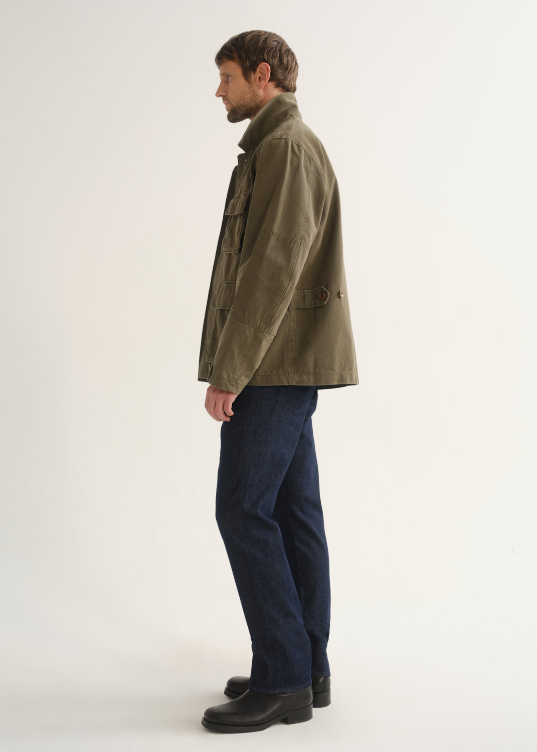 Person wearing dark indigo straight leg jeans with an olive green jacket and black boots, side full body view on a light grey background.