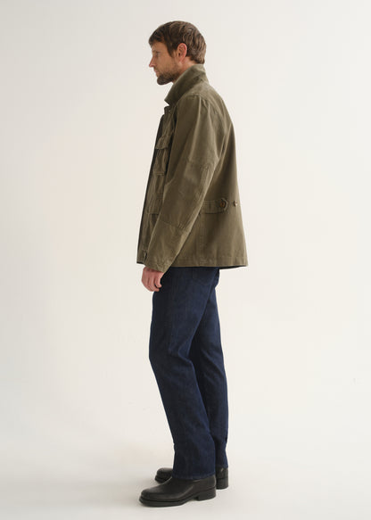 Person wearing dark indigo straight leg jeans with an olive green jacket and black boots, side full body view on a light grey background.