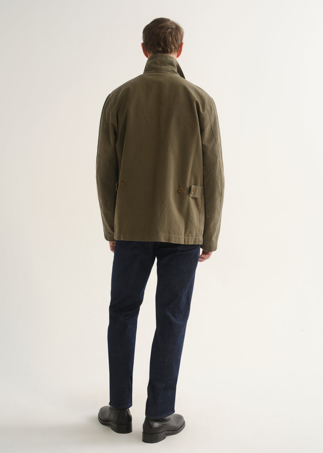 Person wearing dark indigo straight leg jeans with an olive green jacket and black boots, back full body view on a light grey background.