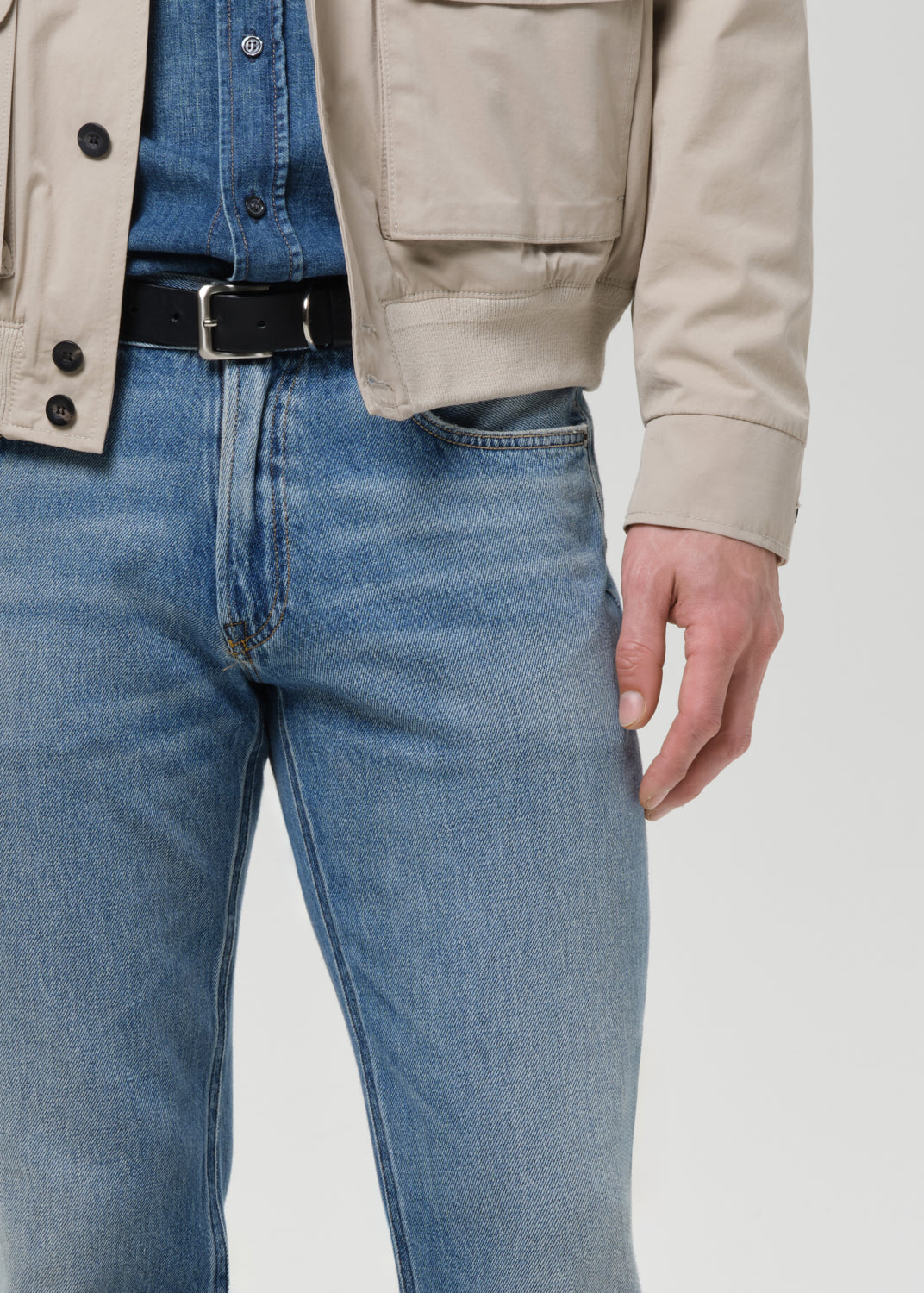 Person wearing a beige jacket, blue jeans, and a black belt on a light gray background