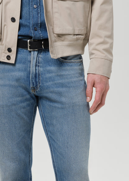 Person wearing a beige jacket, blue jeans, and a black belt on a light gray background