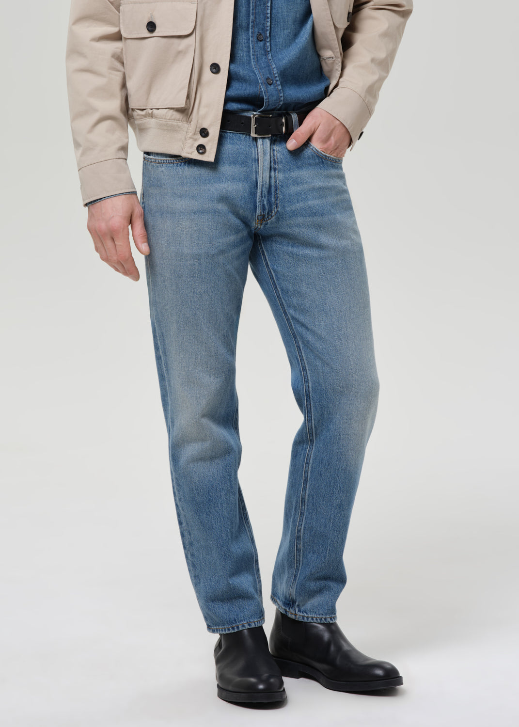 Person wearing blue jeans, a beige jacket, and black shoes on a plain background