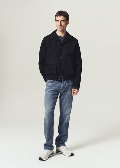 A man modeling a black jacket, blue jeans, and white sneakers against a light gray studio background.