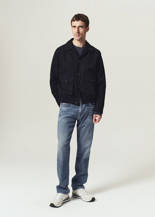 A man modeling a black jacket, blue jeans, and white sneakers against a light gray studio background.