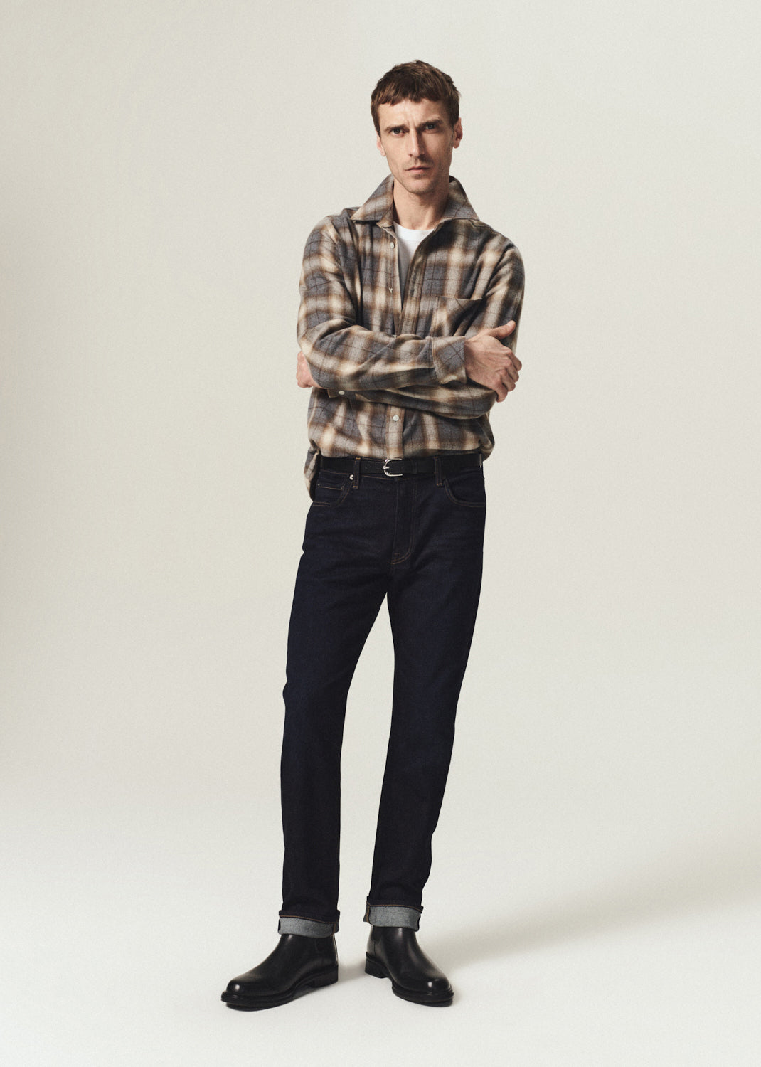 A man modeling a brown and beige plaid button-down shirt, dark blue jeans, and black leather boots against a light gray studio background.