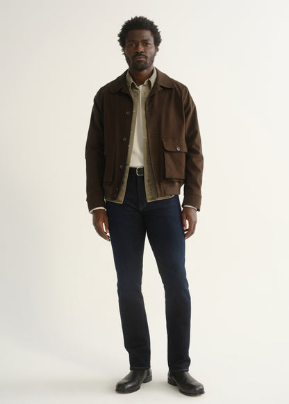 Person wearing dark indigo jeans with a brown jacket, dark shirt, and black shoes, front full body view on a light grey background.