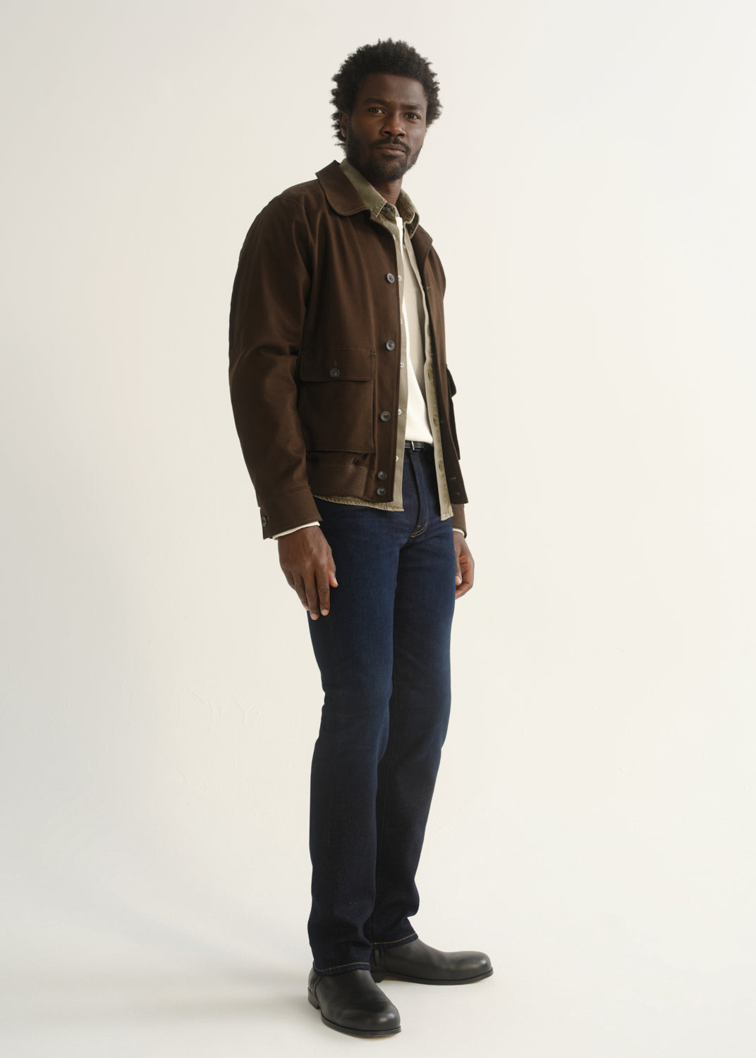 Person wearing dark indigo jeans with a brown jacket and black shoes, side full body view on a light grey background.