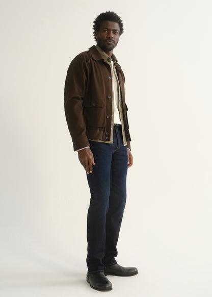 Person wearing dark indigo jeans with a brown jacket and black shoes, side full body view on a light grey background.