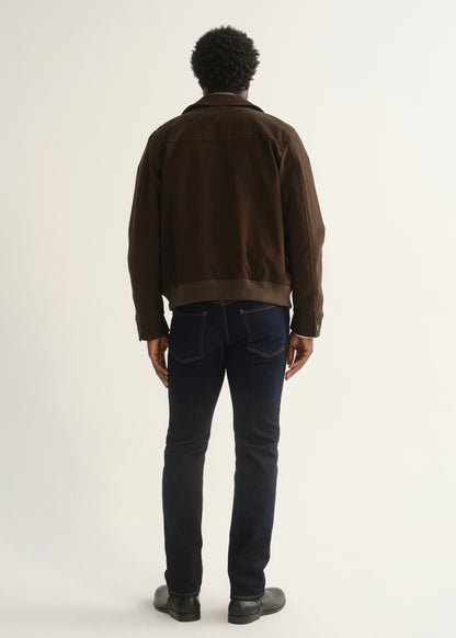 Person wearing dark indigo jeans with a brown jacket and black shoes, back full body view on a light grey background.