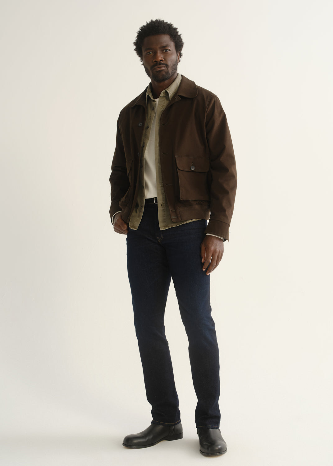 Person wearing dark indigo jeans with a brown jacket, dark shirt, and black shoes, angled front full body view on a light grey background.