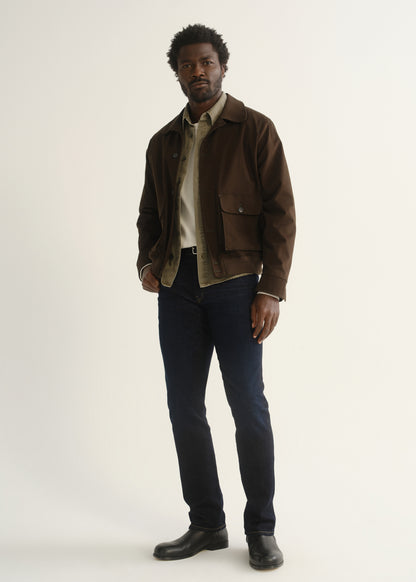 Person wearing dark indigo jeans with a brown jacket, dark shirt, and black shoes, angled front full body view on a light grey background.