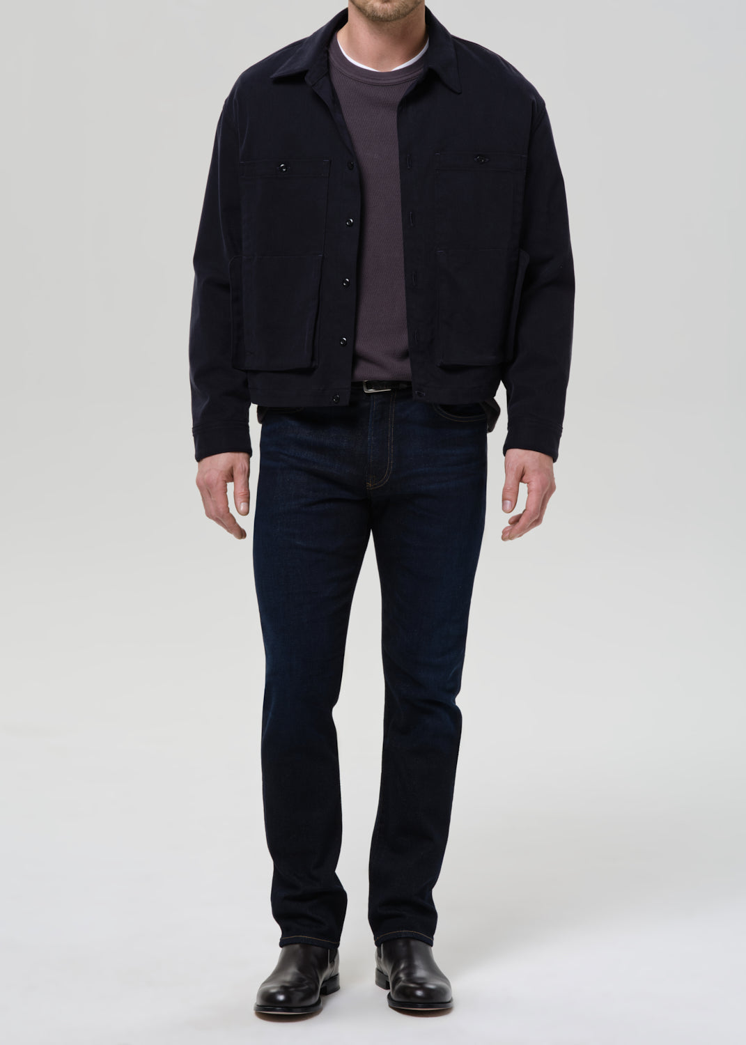 Person wearing dark indigo straight leg jeans with a black bomber jacket and dark charcoal crewneck tee on a grey background.