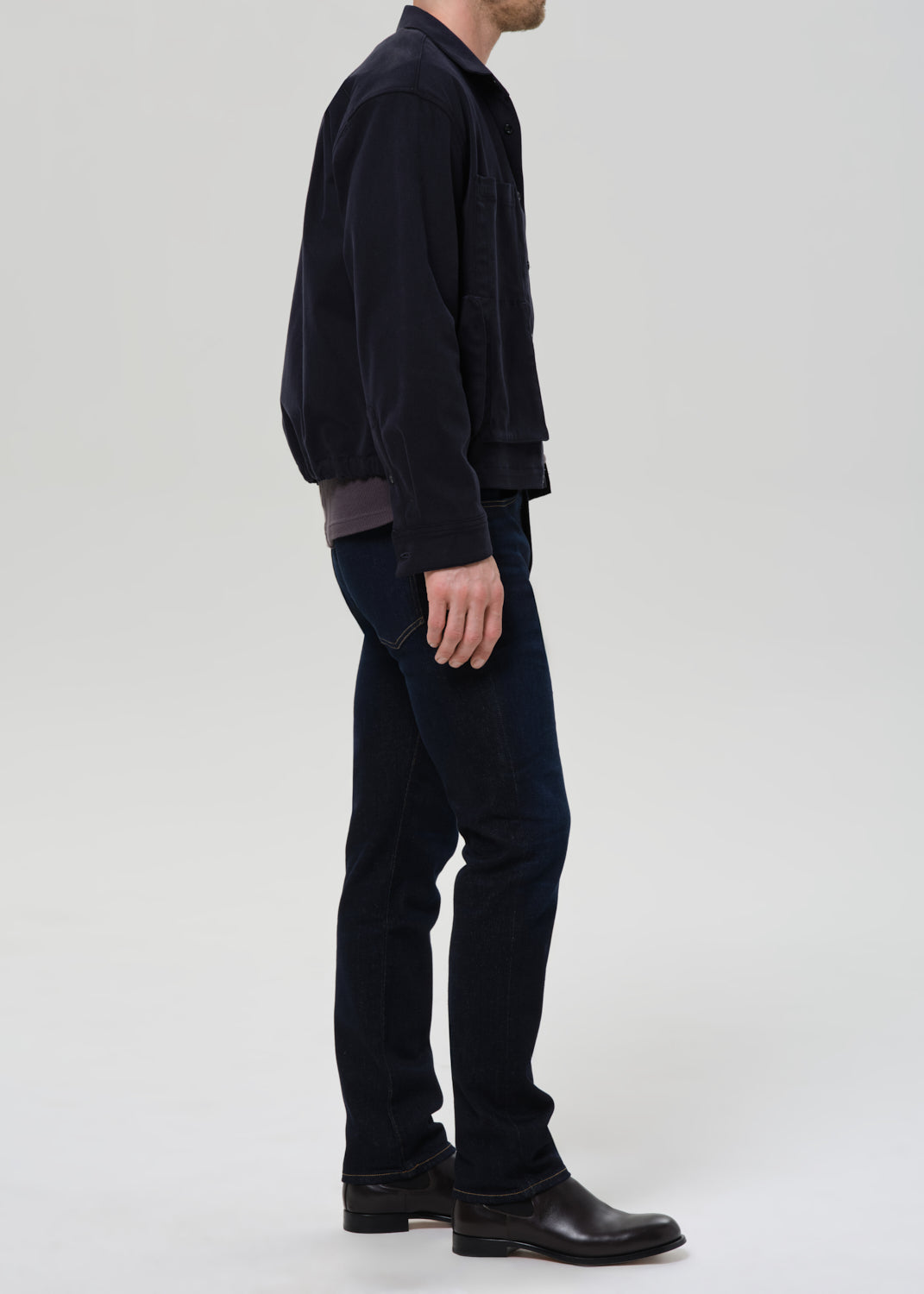 Person wearing dark indigo straight leg jeans with a black bomber jacket and dark charcoal crewneck tee viewed from the side on a grey background.