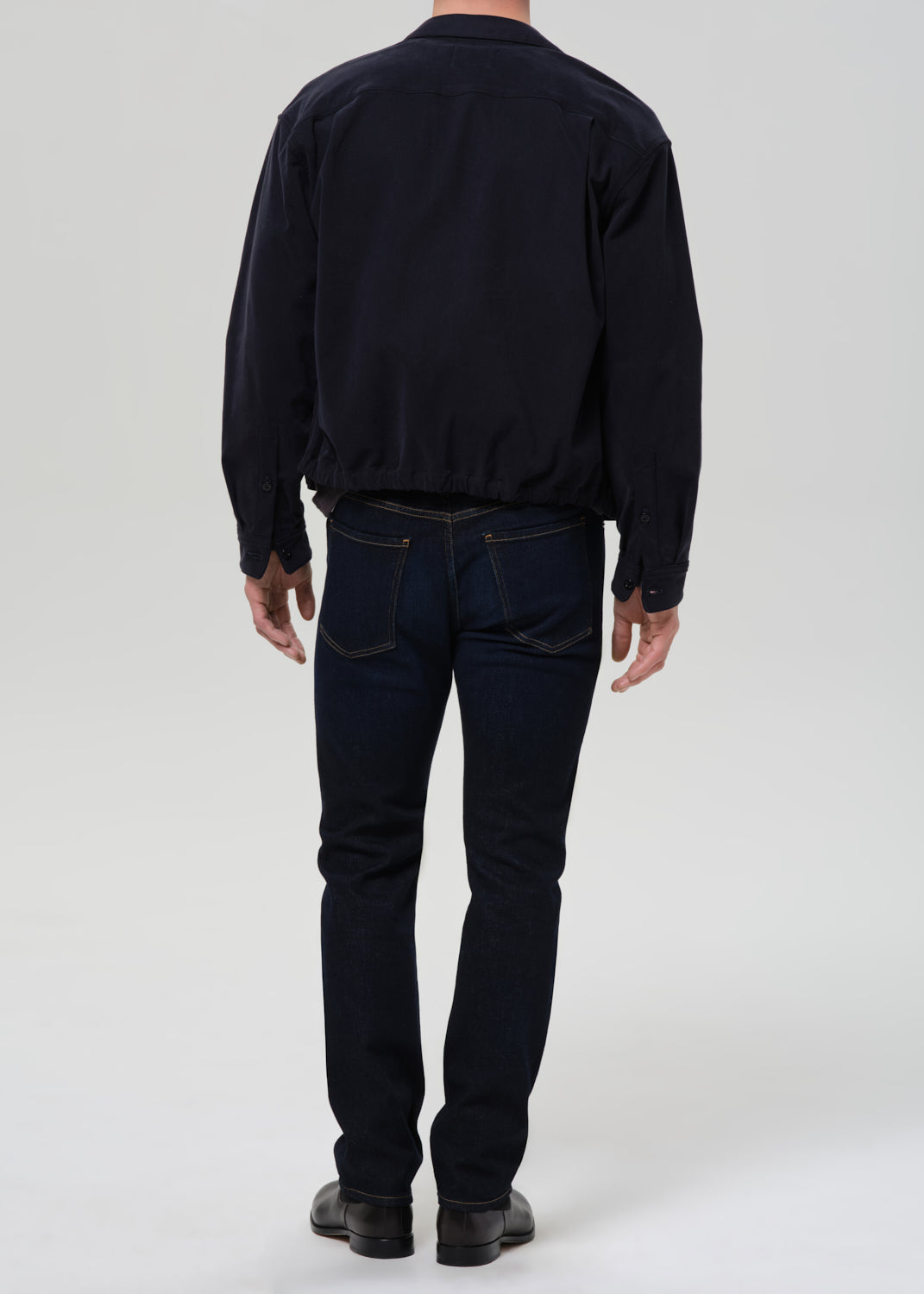 Person wearing dark indigo straight leg jeans with a black bomber jacket and dark charcoal crewneck tee viewed from the back on a grey background.