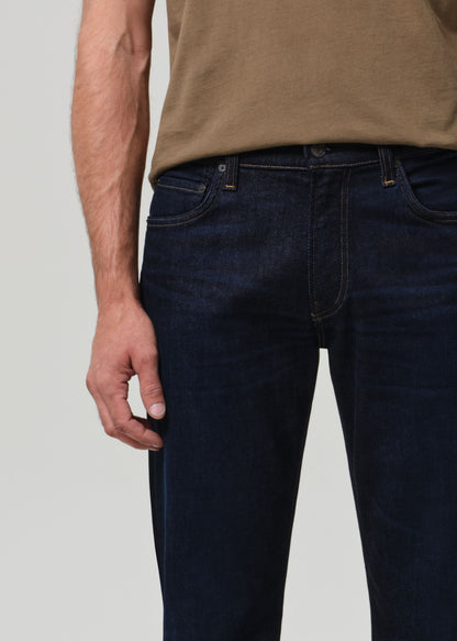 Person wearing dark blue jeans and a brown shirt on a light gray background