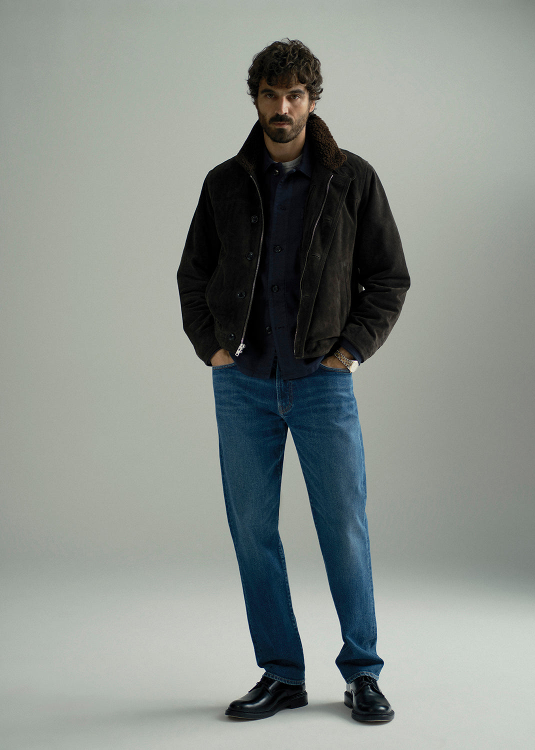 A man stands in a studio wearing a dark brown suede jacket with a brown shearling collar, a navy blue shirt, blue jeans, and black leather shoes against a light gray background.