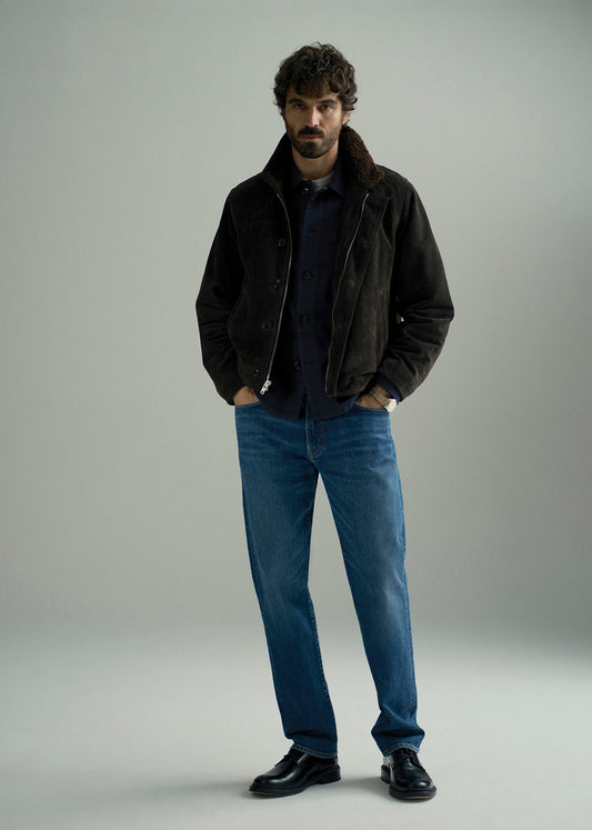 A man stands in a studio wearing a dark brown suede jacket with a brown shearling collar, a navy blue shirt, blue jeans, and black leather shoes against a light gray background.