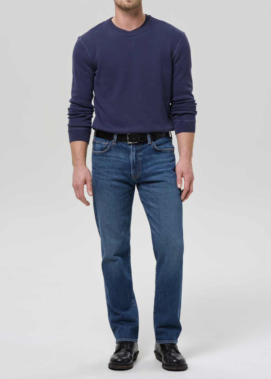A man wearing a navy blue long-sleeve crewneck sweater, blue jeans, a black belt, and black leather shoes, standing against a grey background.