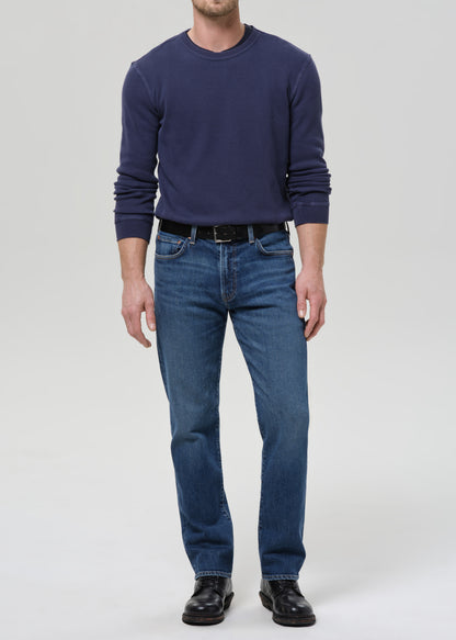 A man wearing a navy blue long-sleeve crewneck sweater, blue jeans, a black belt, and black leather shoes, standing against a grey background.