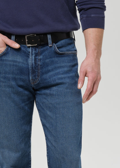 A close-up of a person wearing medium-wash blue jeans with a black belt and a navy ribbed sweater, cropped from the chest down against a grey background.
