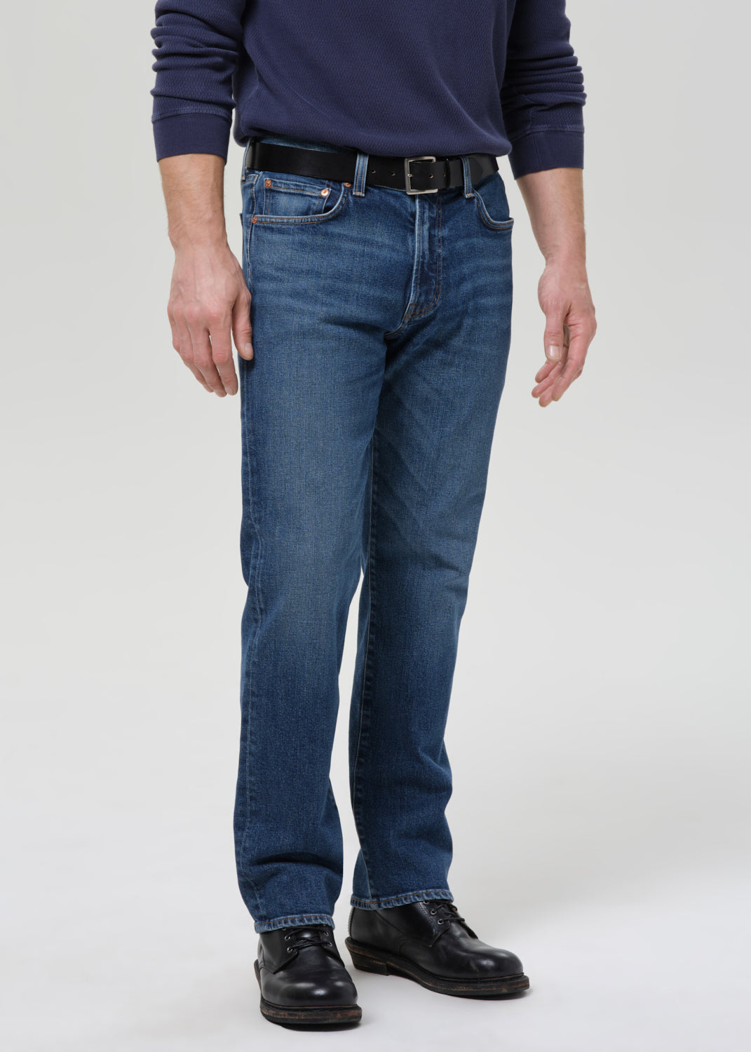 A man wearing a pair of medium-wash blue jeans with a black belt and black leather shoes, shown from the waist down against a grey background.
