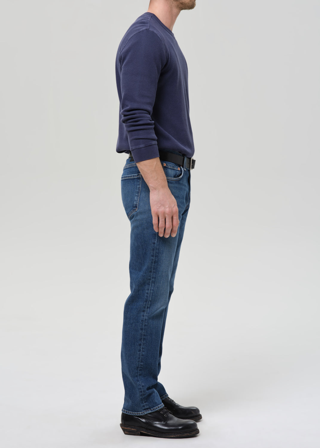 A man shown in profile wearing a navy long-sleeve crewneck sweater, blue jeans, a black belt, and black leather shoes, standing against a grey background.