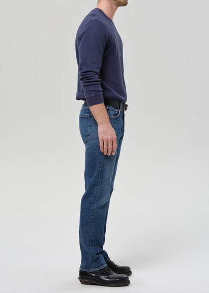 A man shown in profile wearing a navy long-sleeve crewneck sweater, blue jeans, a black belt, and black leather shoes, standing against a grey background.