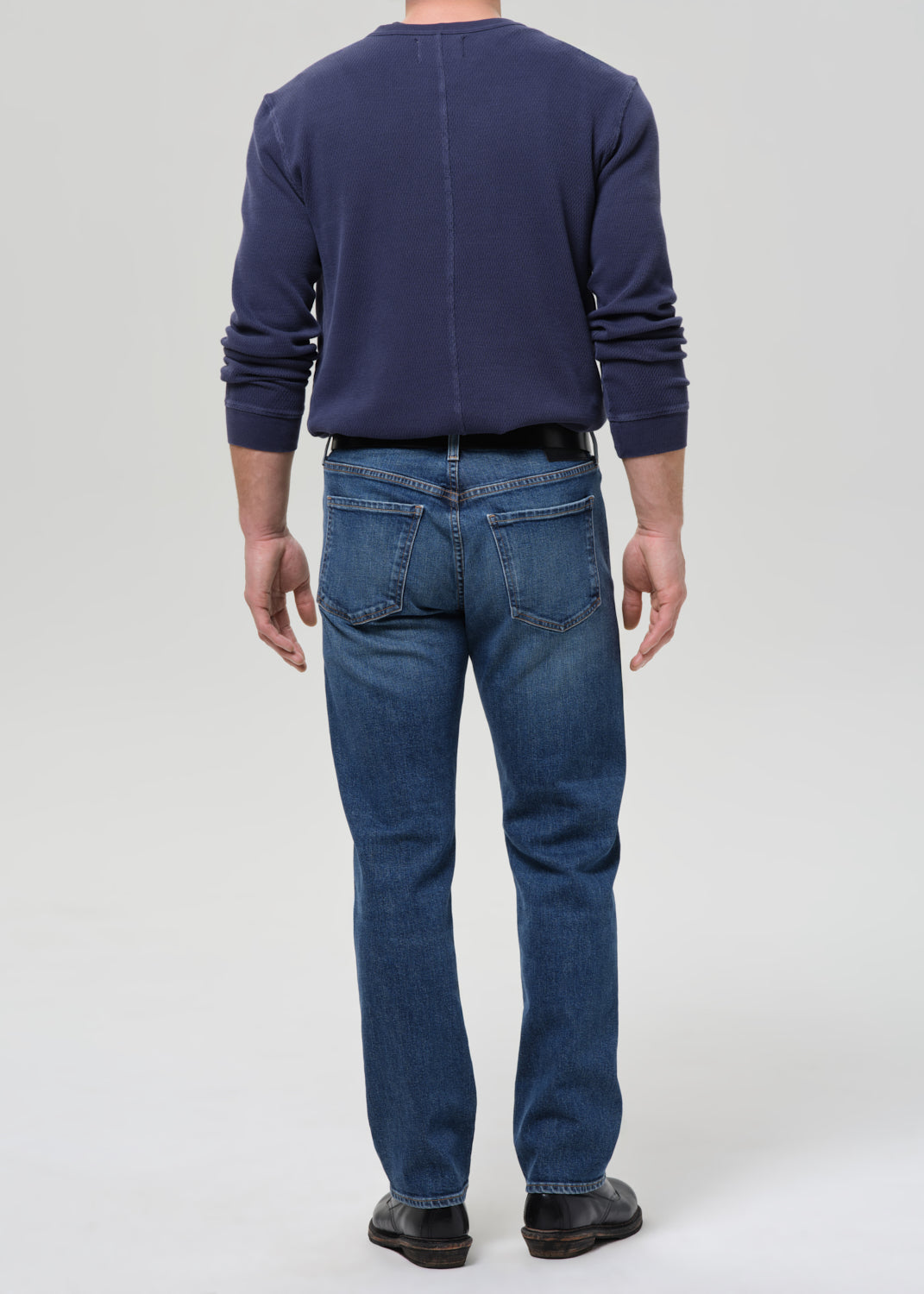 A man viewed from the back wearing a navy blue long-sleeve crewneck sweater, blue jeans, and black shoes, standing against a grey background.