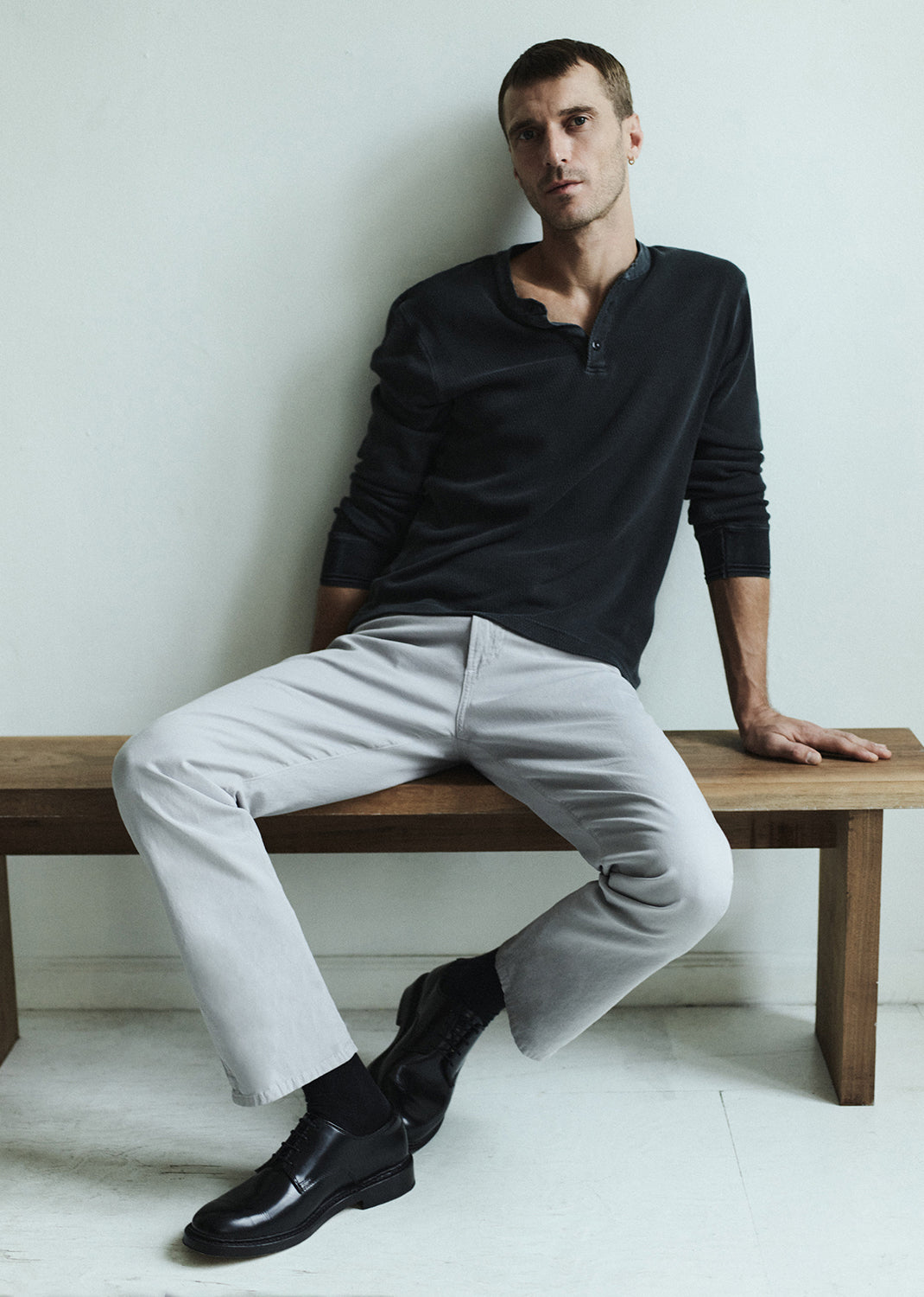 Man sitting on a wooden bench wearing a charcoal long-sleeve shirt, light gray pants, and black shoes.