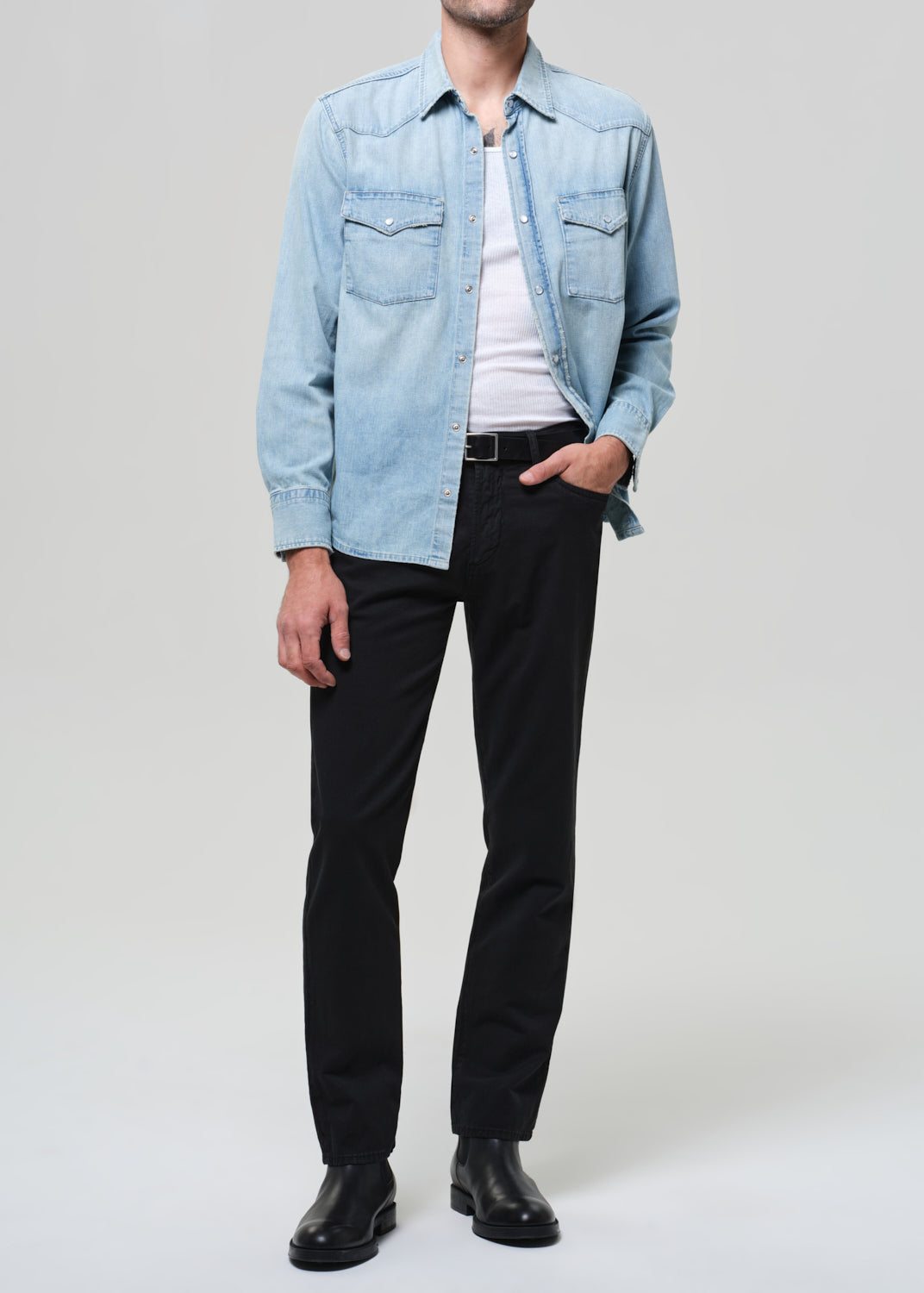 Person wearing a light blue denim shirt, black pants, and black shoes on a plain background