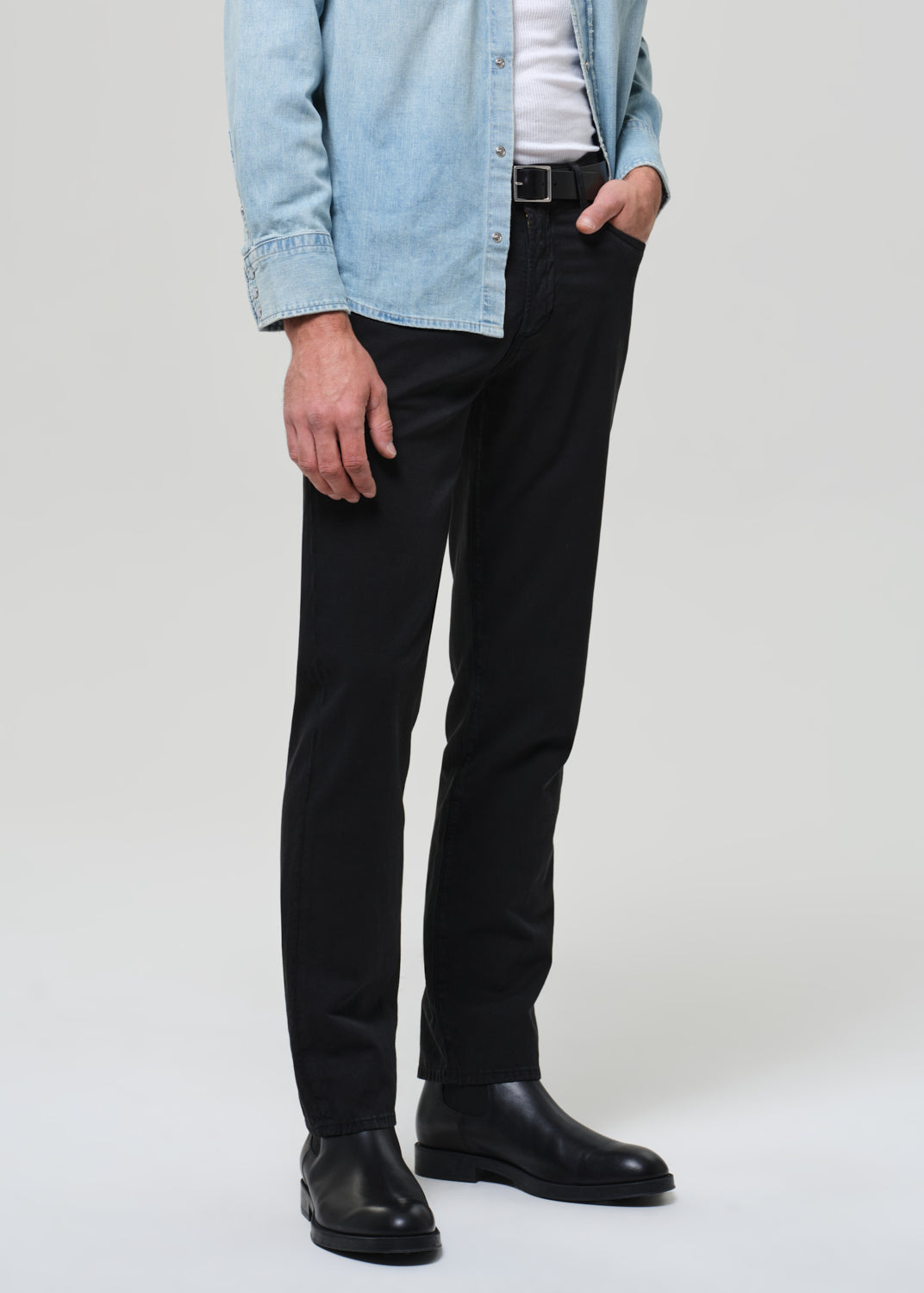 Person wearing a light blue denim shirt, black pants, and black shoes on a plain background