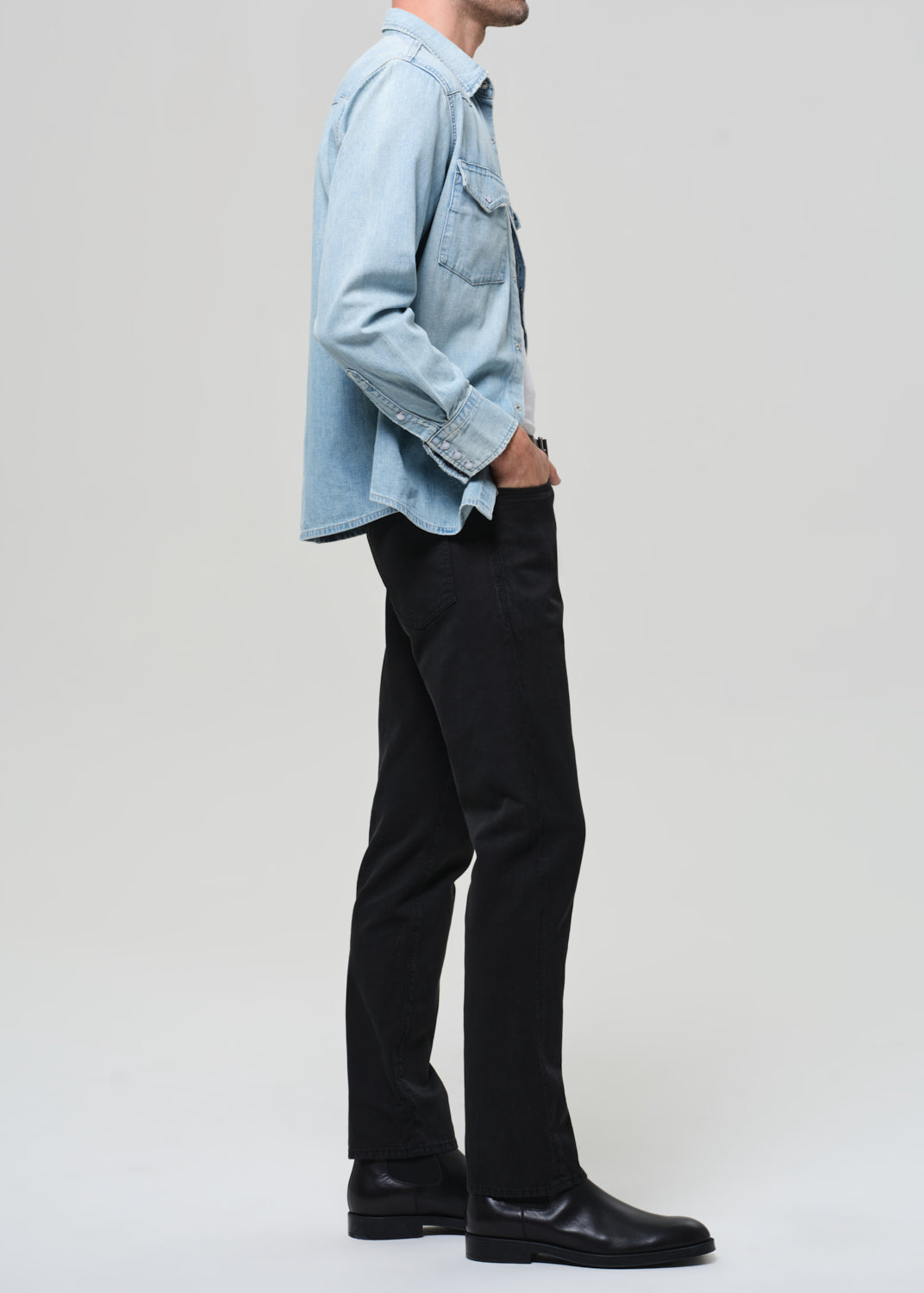Person wearing a light blue denim shirt, black pants, and black shoes on a plain background