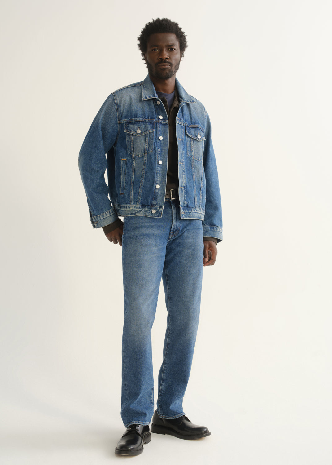 Person wearing medium blue straight leg jeans with a blue denim jacket, dark t-shirt, and black shoes, front full body view on a light grey background.