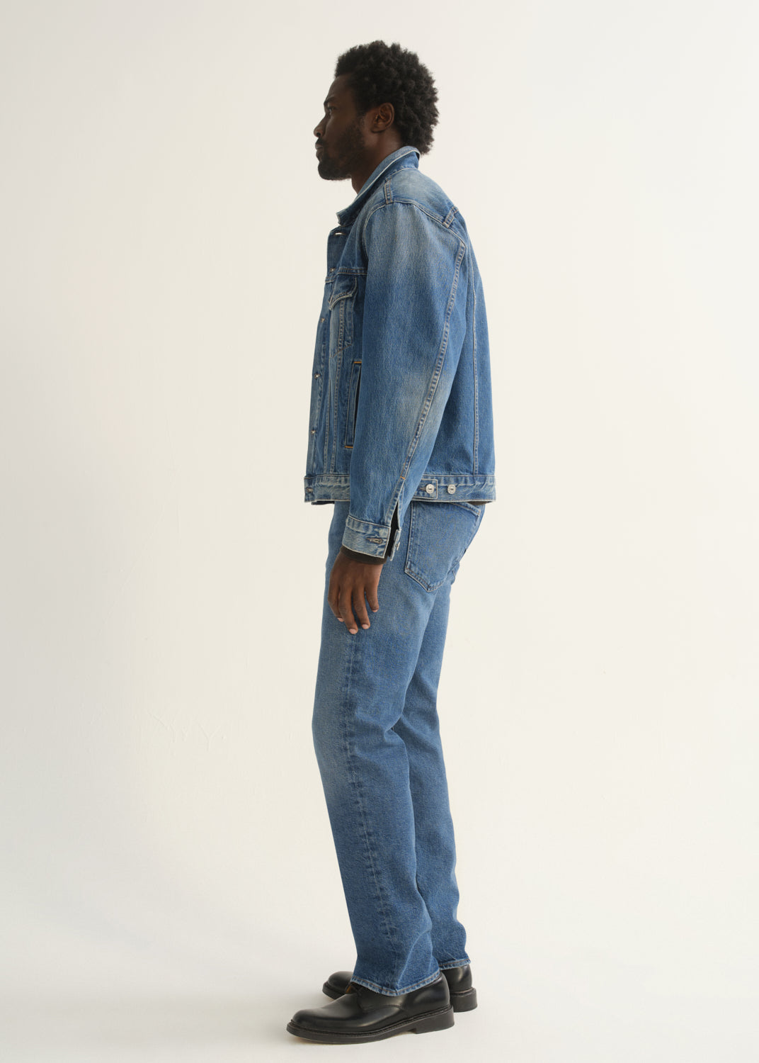 Person wearing medium blue straight leg jeans with a blue denim jacket and black shoes, side full body view on a light grey background.
