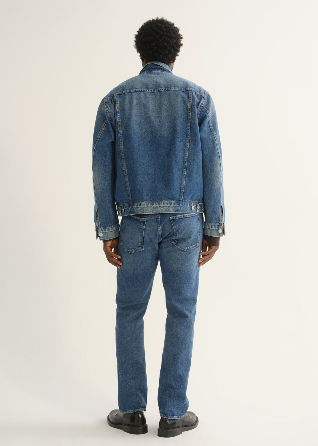 Person wearing medium blue straight leg jeans with a blue denim jacket and black shoes, back full body view on a light grey background.