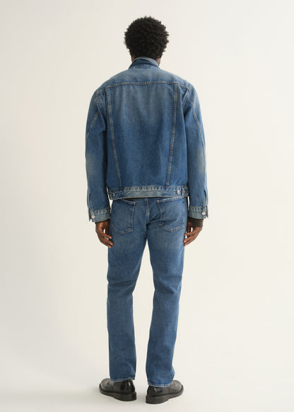 Person wearing medium blue straight leg jeans with a blue denim jacket and black shoes, back full body view on a light grey background.