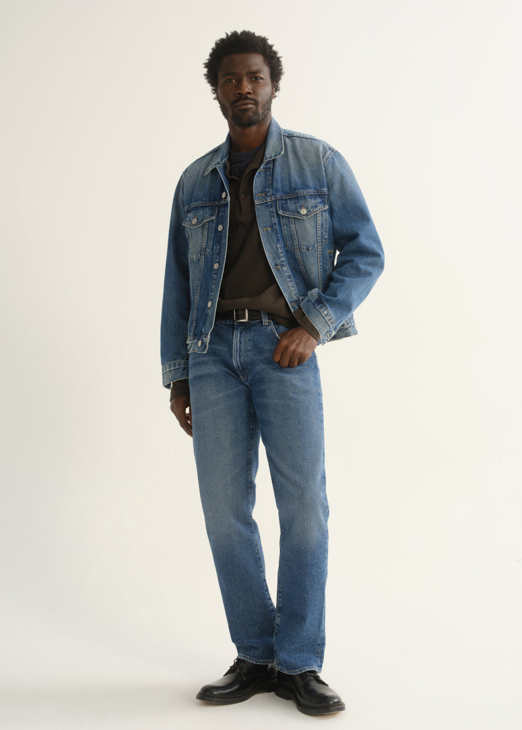 Person wearing medium blue straight leg jeans with a blue denim jacket, dark t-shirt, and black shoes, angled front full body view on a light grey background.