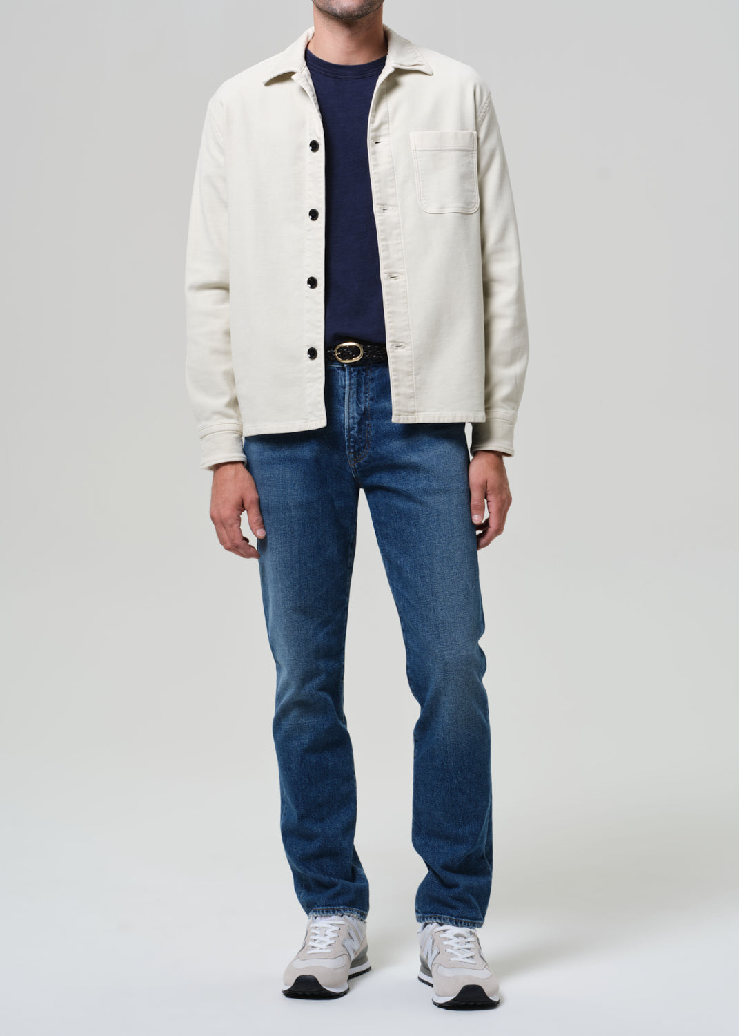 Person wearing a white jacket, navy shirt, blue jeans, and white sneakers on a light gray background