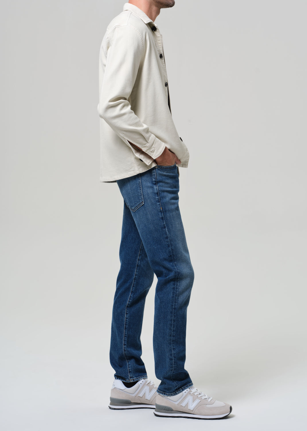 Person wearing a light-colored shirt, blue jeans, and white sneakers on a plain background