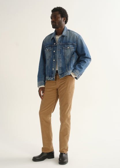 Person wearing camel brown slim straight leg pants with a medium blue denim jacket and black shoes, angled front full body view on a light grey background.