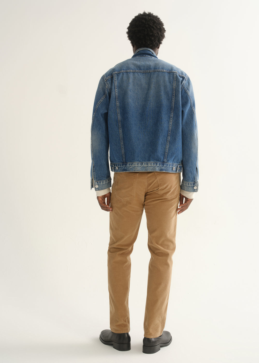 Person wearing camel brown slim straight leg pants with a medium blue denim jacket and black shoes, back full body view on a light grey background.