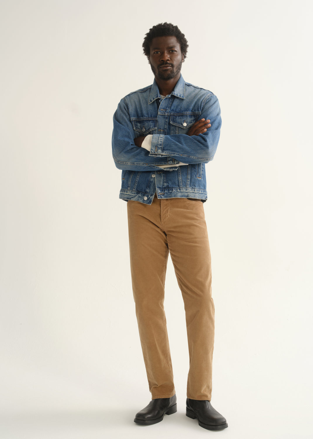 Person wearing camel brown slim straight leg pants with a medium blue denim jacket and black shoes, front full body view with arms crossed on a light grey background.